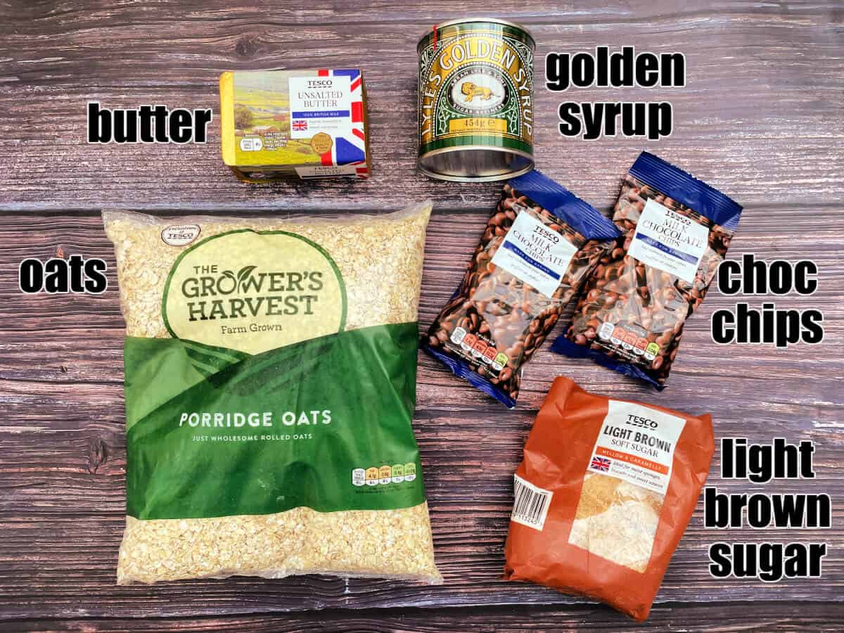Labelled ingredients for dish on a wooden background - oats, butter, golden syrup, choc chips, light brown sugar.