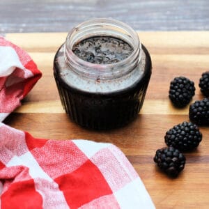 Jar of blackberry jam, red checked dish towel and blackberries to the sides.