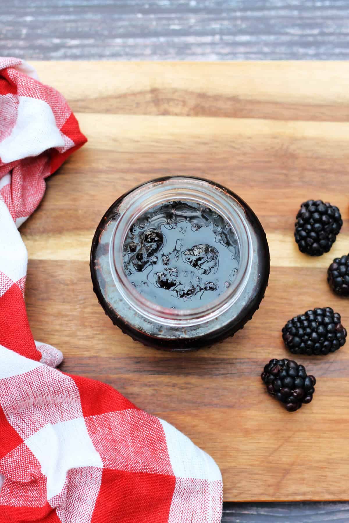 Open jar of jam on wooden board with blackberries and checked dish towel, from overhead.