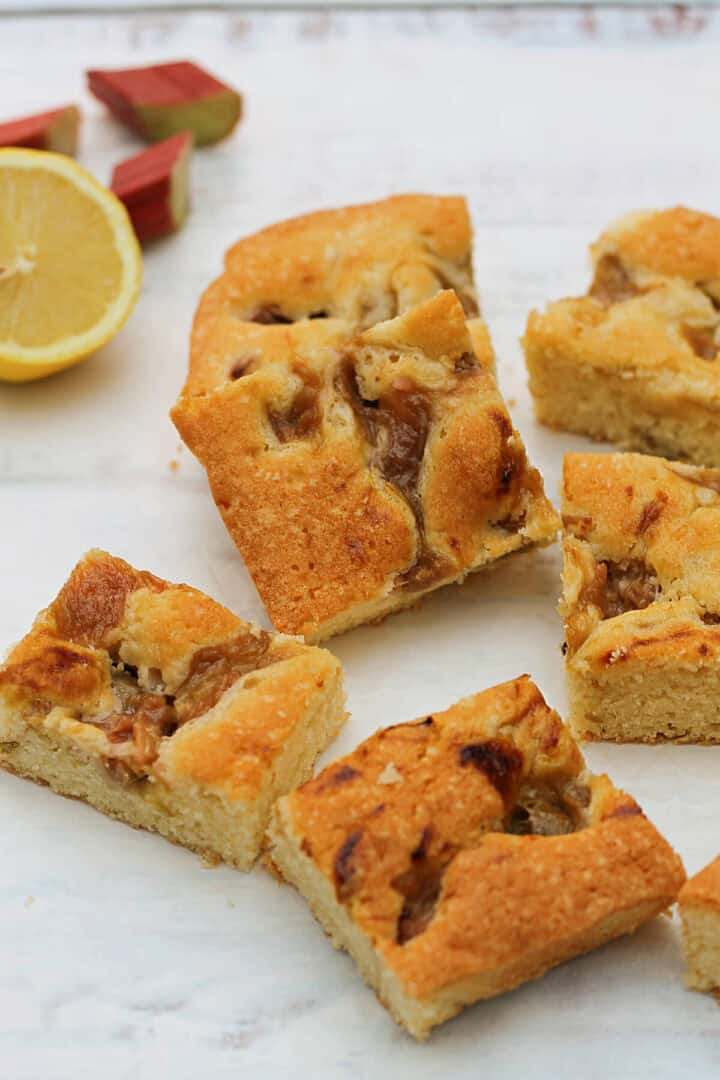 Squares of rhubarb cake on white background, lemon and rhubarb pieces in background.