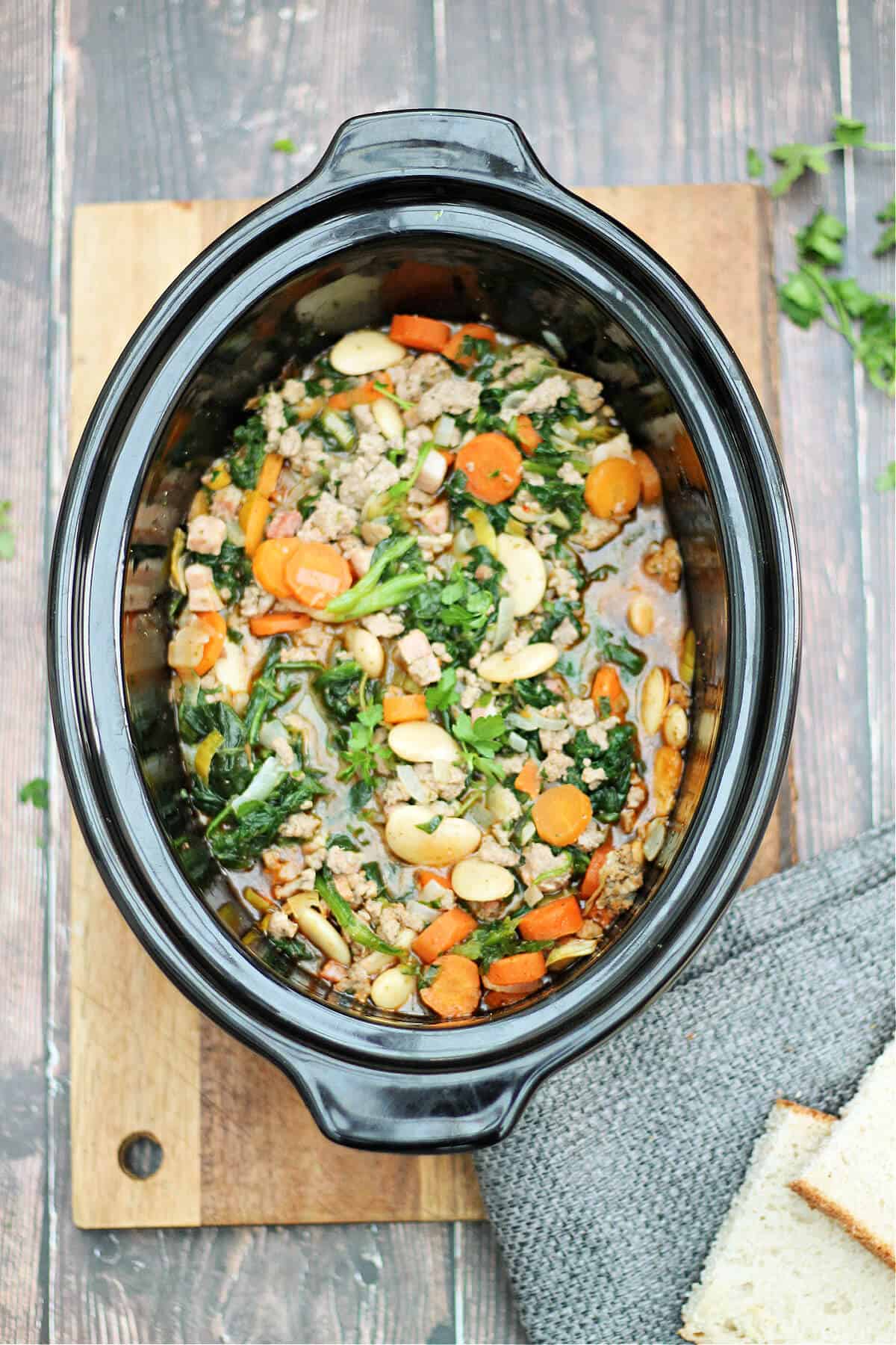 Slow cooker pot with turkey stew with vegetables in it, bread and herbs to the side.