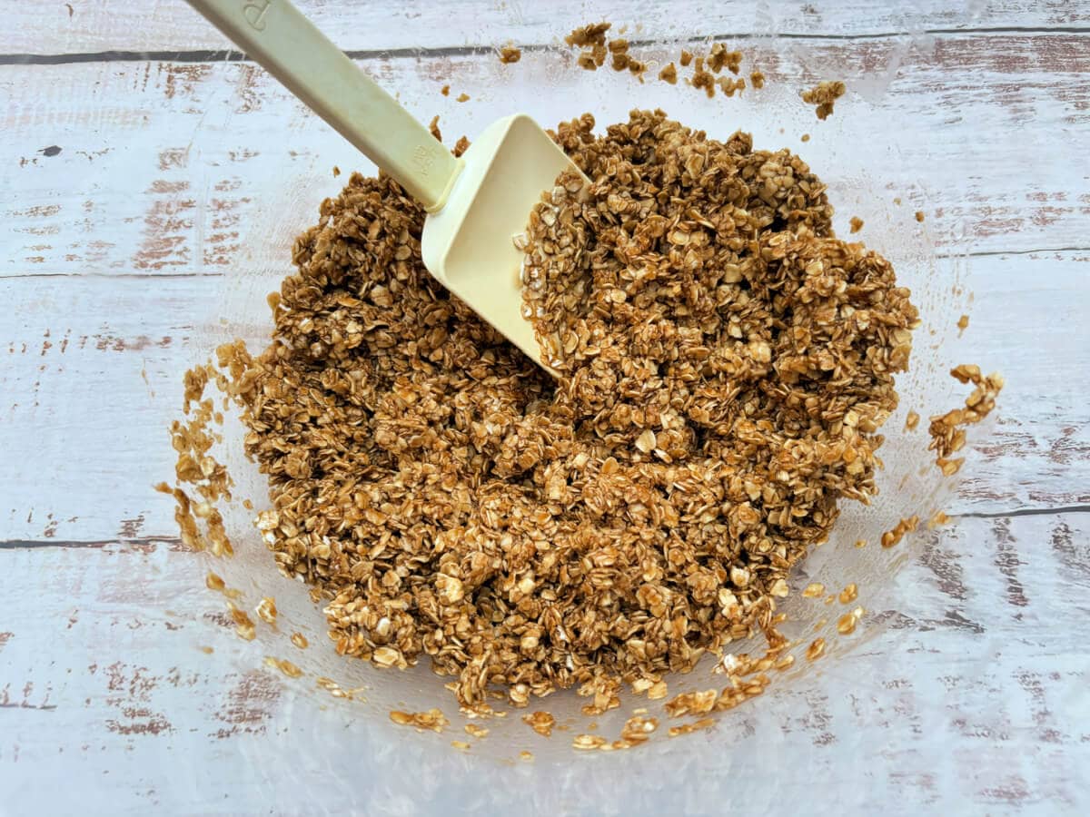 A clear bowl filled with oats coated with the melted butter and sugar mixture, with a spatula.