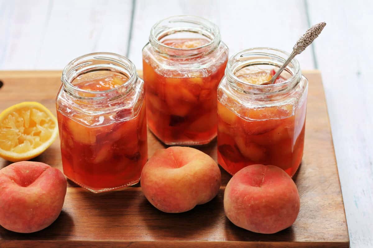 Jar of jams on a board, with peaches and lemon to the side.