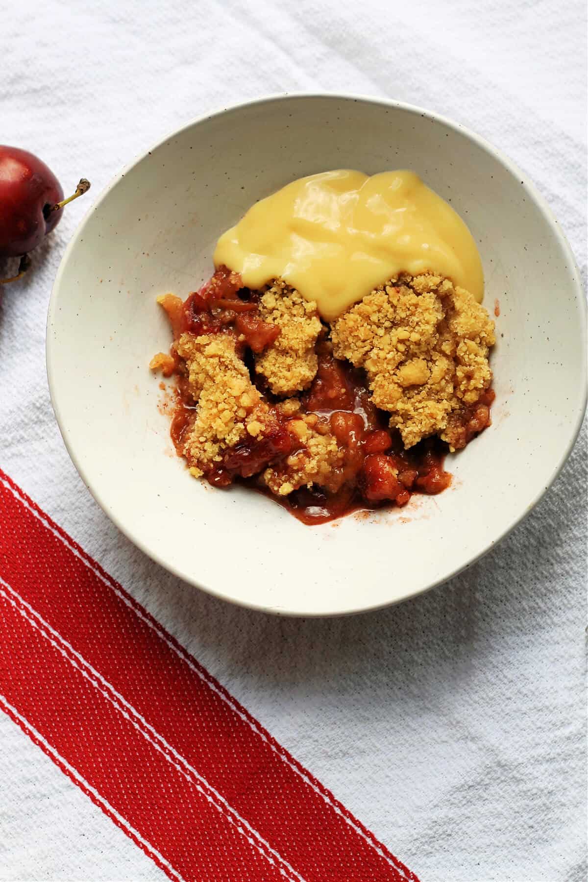 Bowl of plum crumble with custard on a white and red cloth.