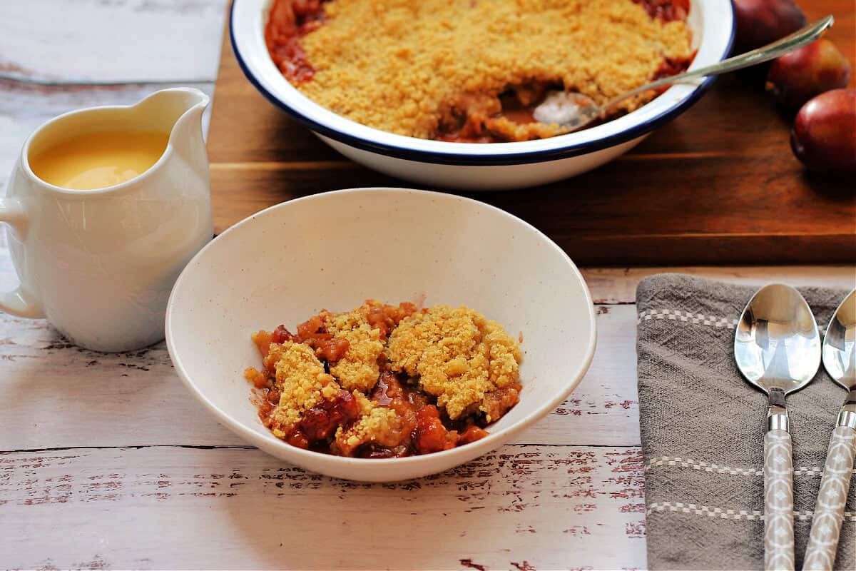 Serving dish of crumble with a separate bowl of crumble and custard jug.