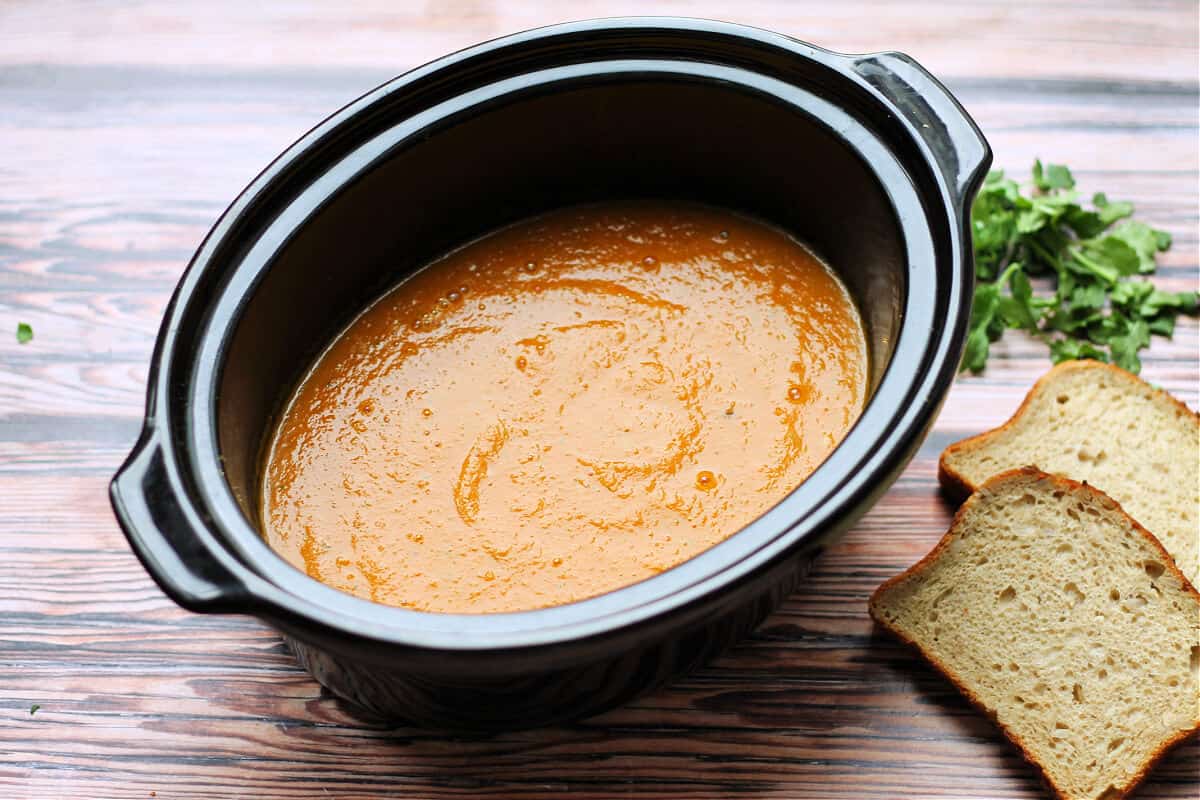 Slow cooker pot containing carrot soup, with brown bread and coriander/cilantro to the side.