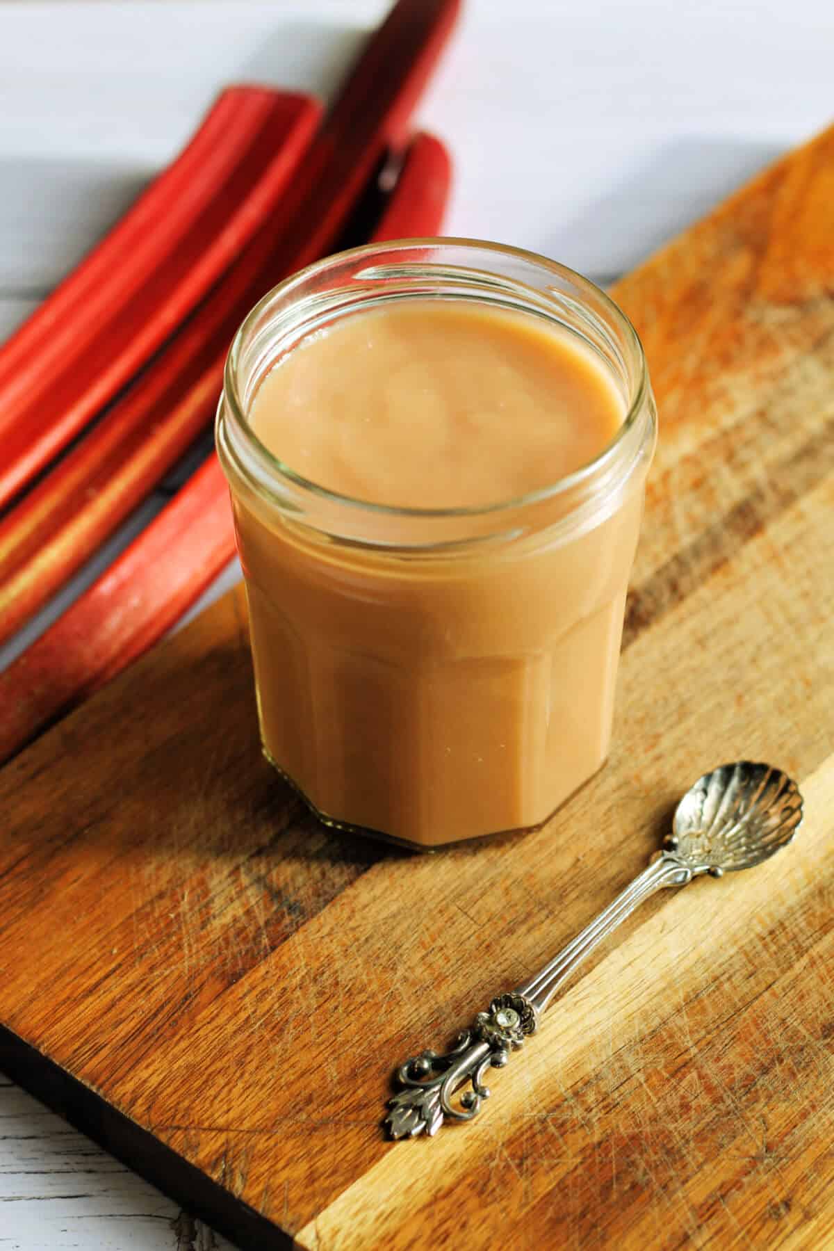 Jar of curd on a wooden board with spoon next to it and rhubarb stalks in background.