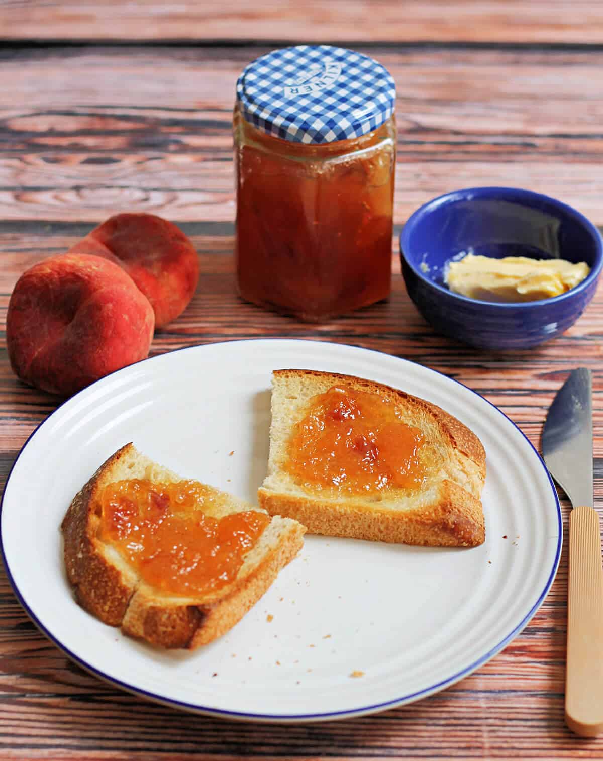 Peach jam on toast on white plate, jar of jam, butter and peaches in background.